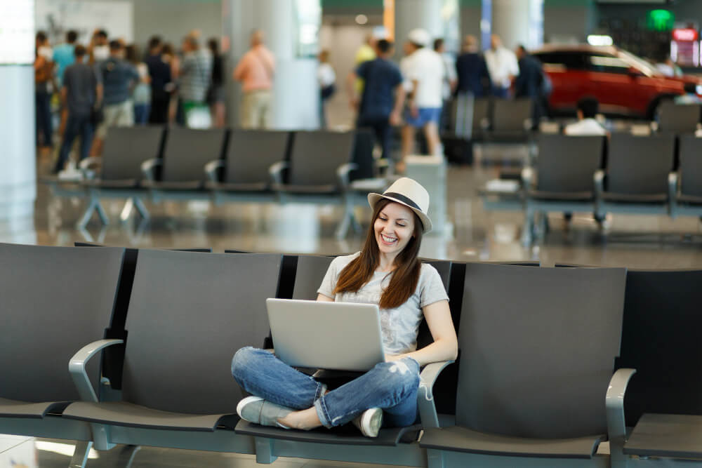 young laughing traveler tourist woman hat sitting with crossed legs working laptop wait lobby hall international airport 1
